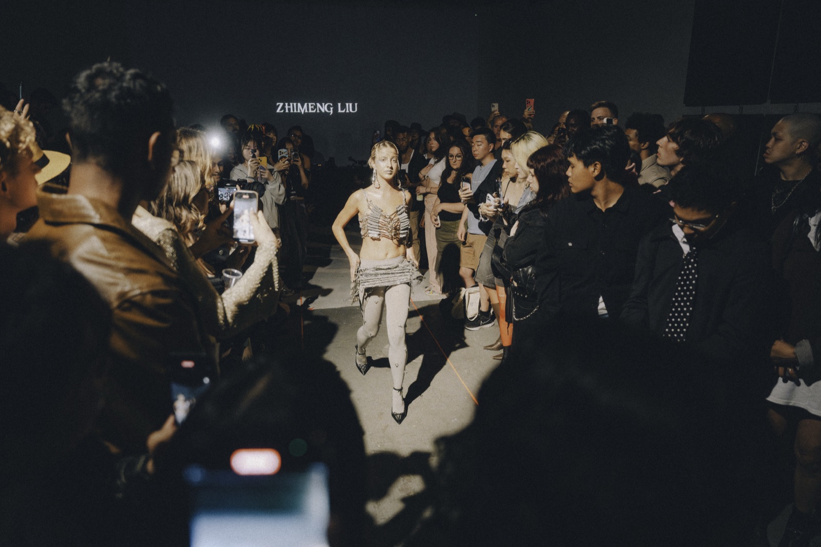 Seen from inside the crowd, a model walks the open floor under a designer's name on the back wall.
