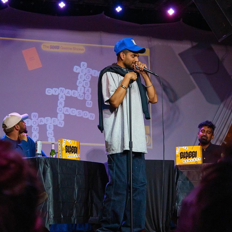 A comedian pitches into a microphone on a purple-lit stage in front of the projected game board.