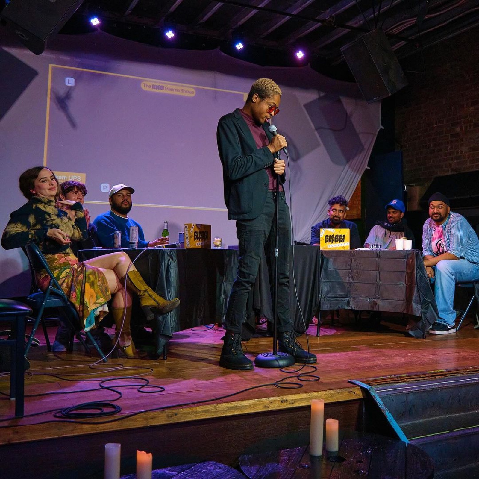 A contestant in a dark suit pitches at the center mic while players at both team tables react.
