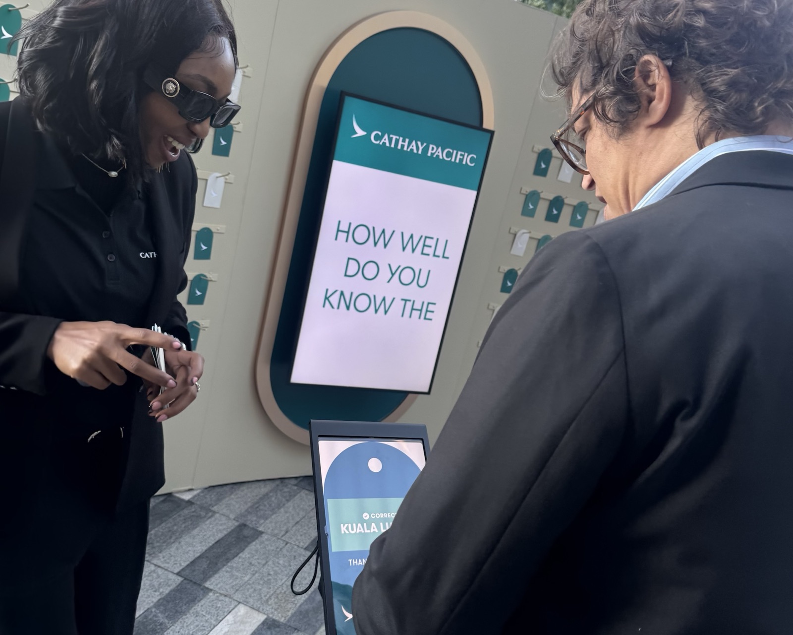 A guest smiles at a vertical screen in an arched frame reading How well do you know the..., next to a tablet kiosk.