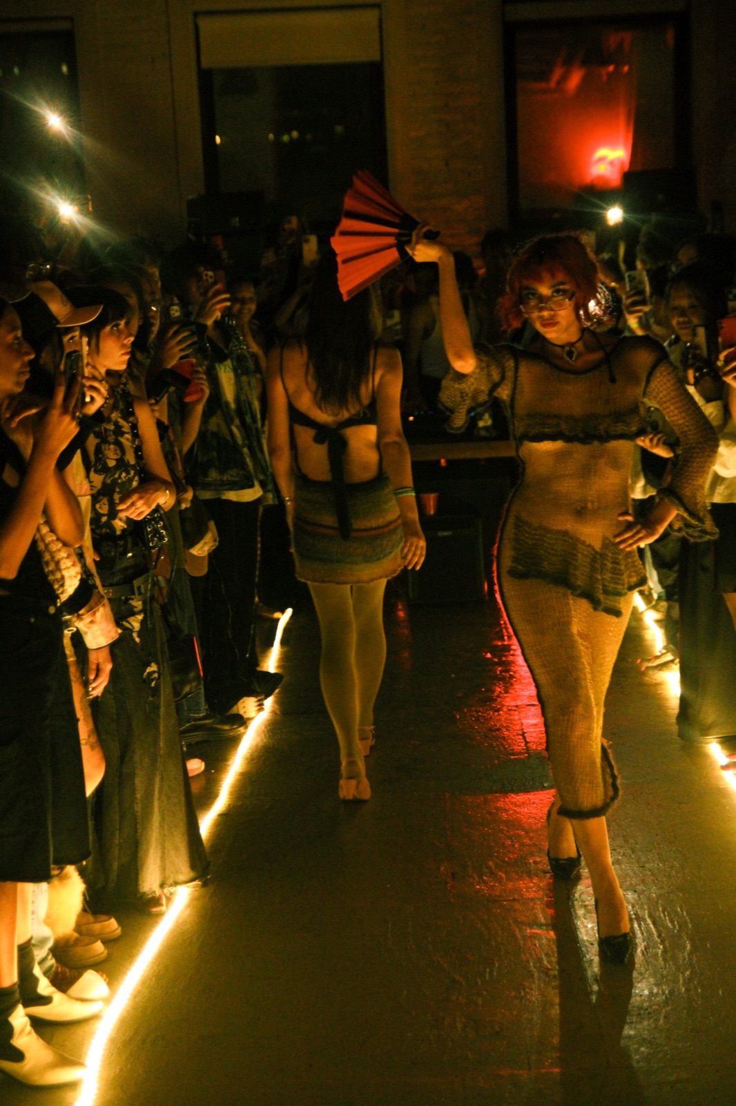 Models in dark looks walk the lit runway through a crowd in near darkness.