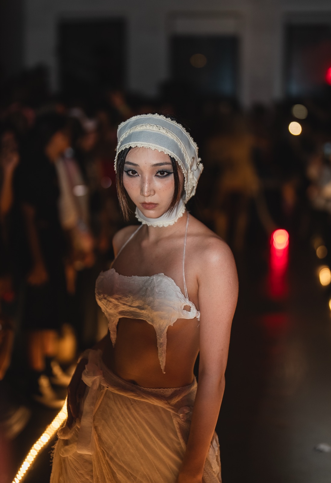 A model in a draped top and knit hood stands on the dark floor with red lights behind them.