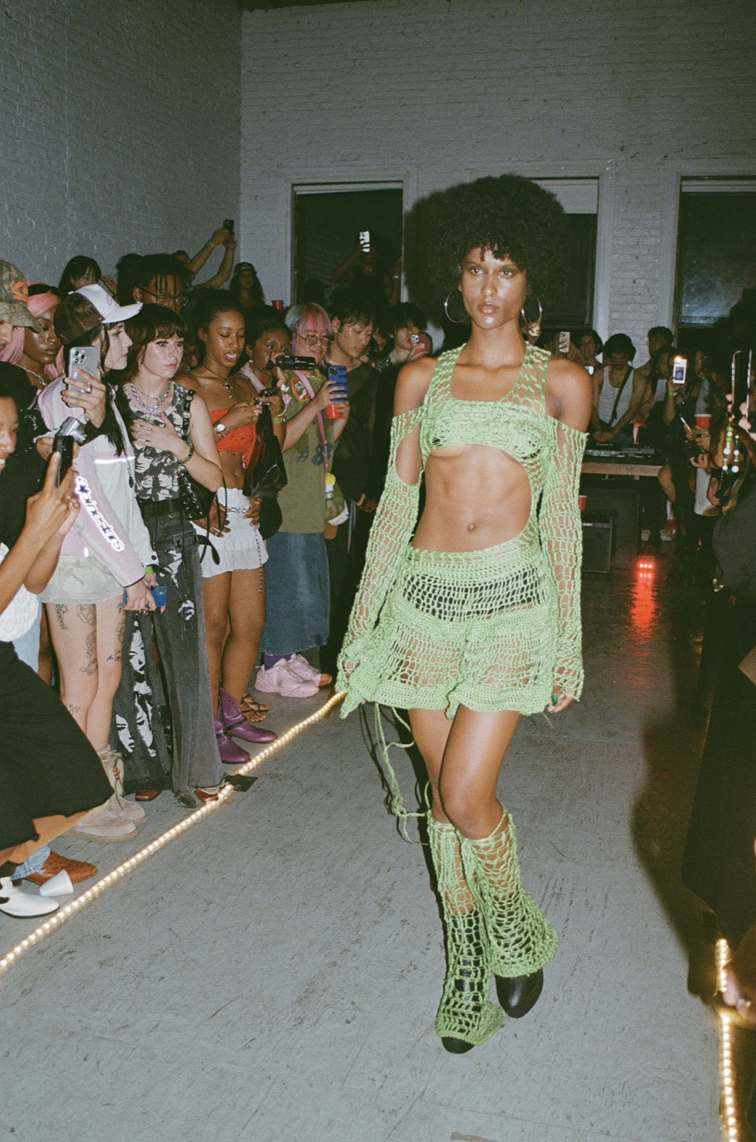 On film: a model in a green crochet set and lace-up boots walks past a line of people watching.