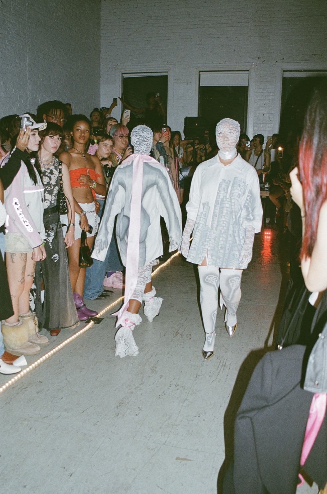 On film: two models in white looks that cover their faces walk side by side down the runway.